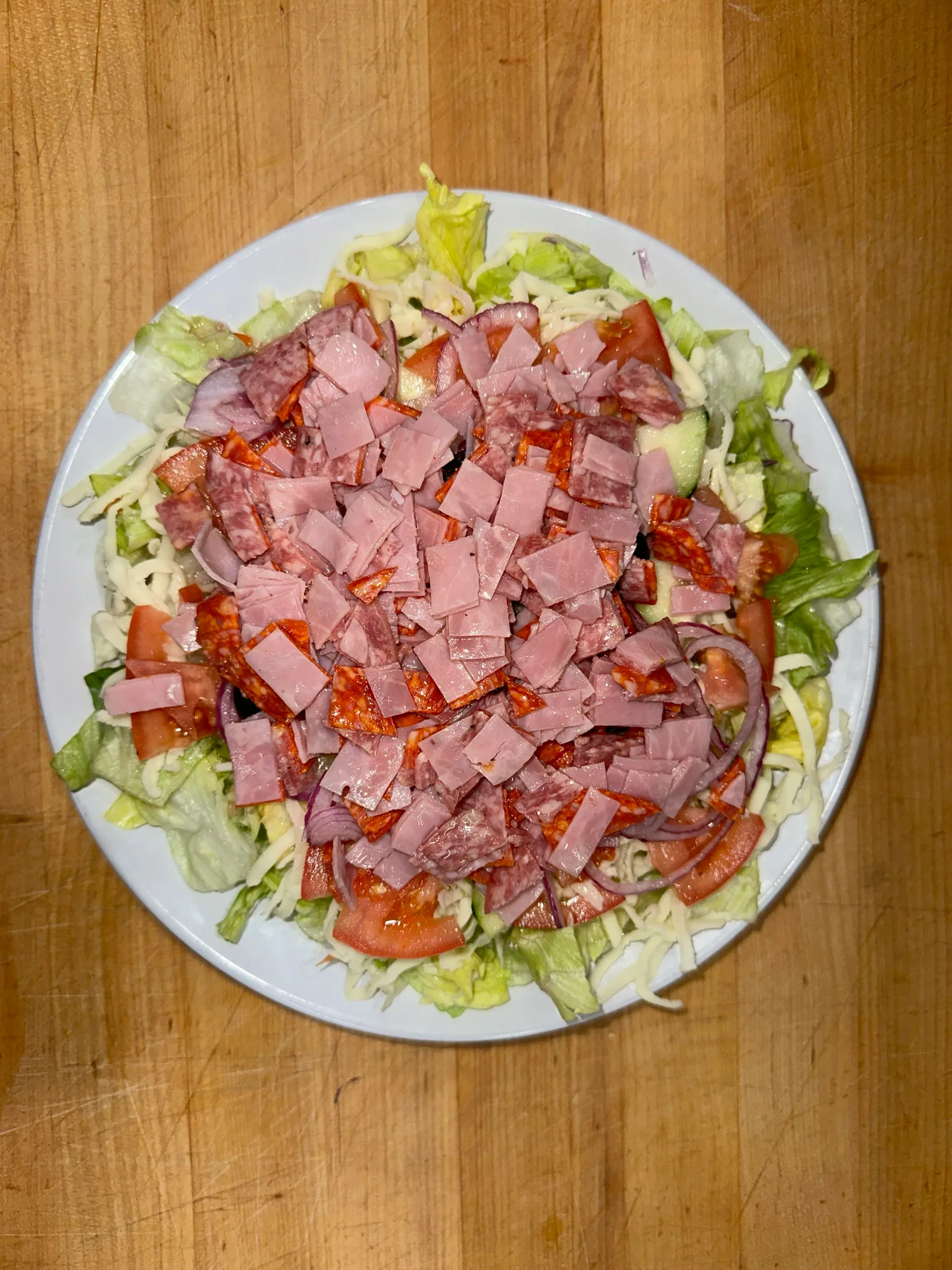 Plate of salad with lettuce, tomatoes, onions, shredded cheese, and chopped ham on top, placed on a wooden surface.