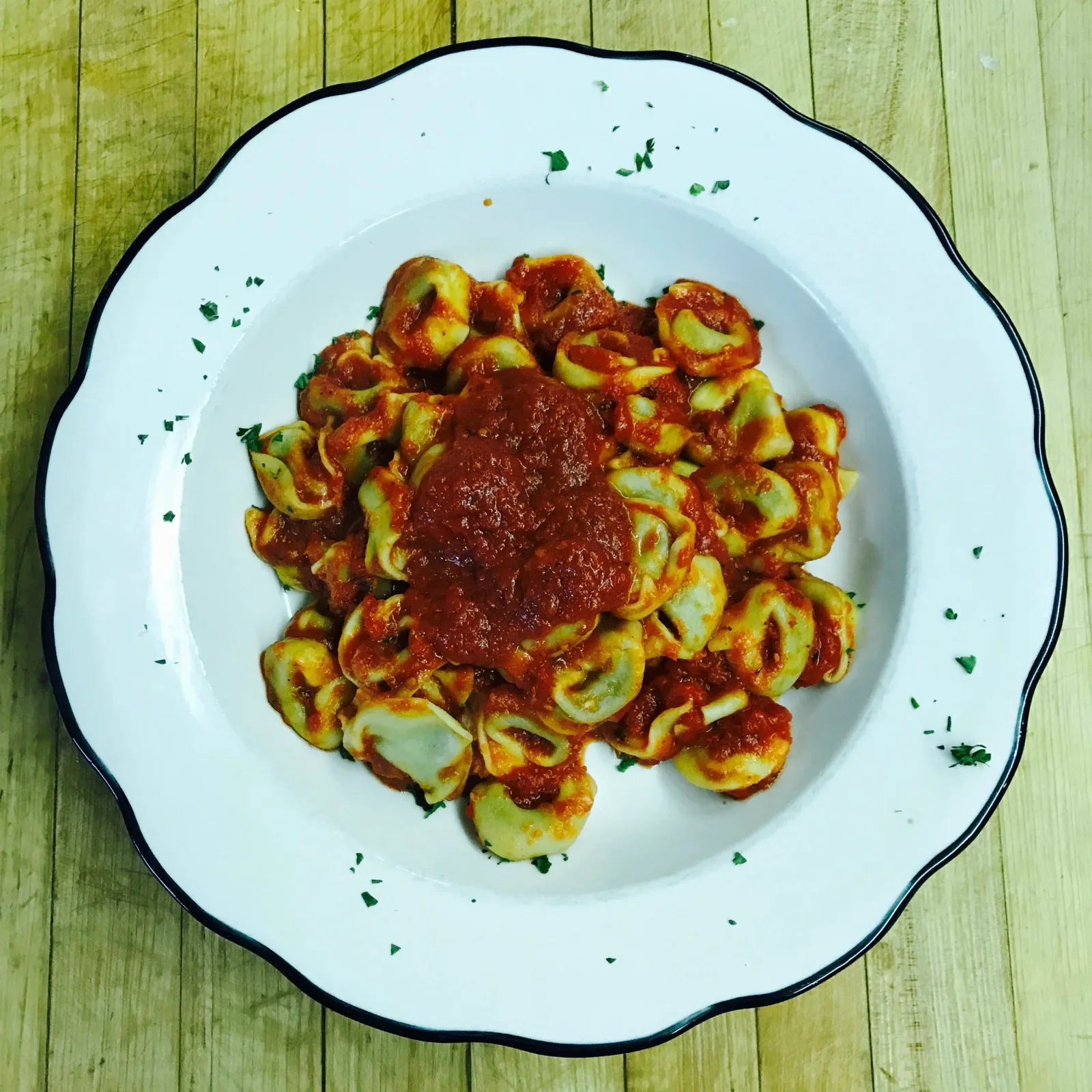 Plate of cooked tortellini pasta with tomato sauce and sprinkled parsley, on a white plate with a black rim on a wooden surface.